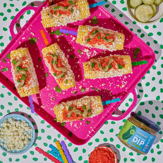 Overhead flat-lay of six corn on the cob sticks topped with red salsa, pickle dip, cotija cheese, and sour cream, arranged on a bright pink tray. Surrounding props include pickles, cheese, dip jars, and a green-and-white polka dot tablecloth.