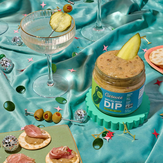 A styled party scene with multiple jars of Dill Pickle Dip, cocktail glasses with pickle garnishes, olives, crackers, and a shiny turquoise satin fabric backdrop.