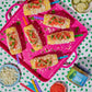 Overhead flat-lay of six corn on the cob sticks topped with red salsa, pickle dip, cotija cheese, and sour cream, arranged on a bright pink tray. Surrounding props include pickles, cheese, dip jars, and a green-and-white polka dot tablecloth.