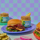 Two jars of Spicy Dill Pickle Dip sandwich spread styled on a table with cheeseburgers and pickles, set against a pastel purple and light blue checkered background.