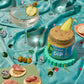 A styled party scene with multiple jars of Dill Pickle Dip, cocktail glasses with pickle garnishes, olives, crackers, and a shiny turquoise satin fabric backdrop.